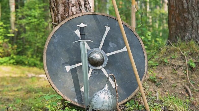 A shield and helmet are placed against a tree in a forest in Eastern Europe. The equipment reflects 11th-century design and craftsmanship, capturing the essence of medieval life