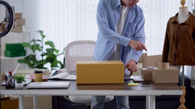 Asian male entrepreneur sealing yellow cardboard box with tape at desk in home business office workspace