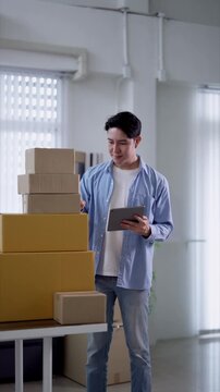 Young Asian entrepreneur checking tablet while managing inventory of stacked shipping boxes in modern warehouse office