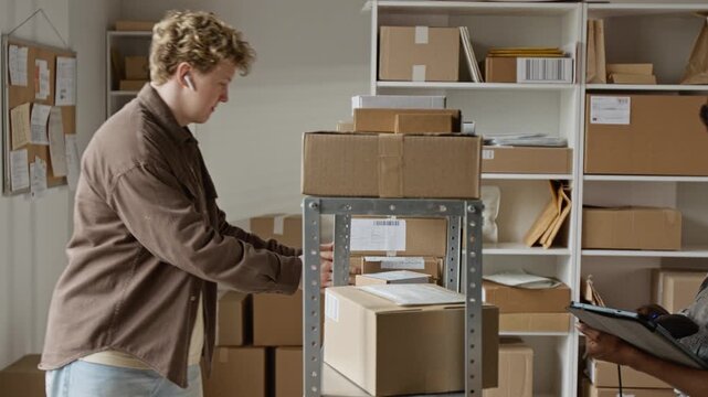 Caucasian young adult man placing cardboard boxes on shelves while African American young adult woman scanning barcodes using tablet. Perfect for e-commerce logistics and retail supply chain ads