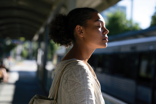 A young woman stands peacefully at a train station, with closed eyes and a serene expression, symbolizing calmness amidst the hustle of urban life and transportation.