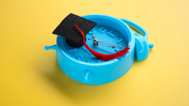 Graduation hat placed on blue clock on bright yellow background concept for academic study time management and deadlines