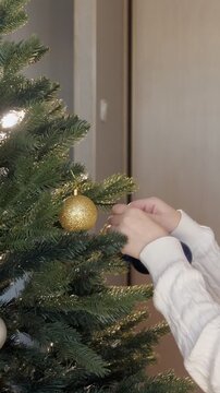 Close up of a young woman's hands decorating a christmas tree with colorful ornaments, hanging a classic blue ball on a branch in preparation for the winter holidays