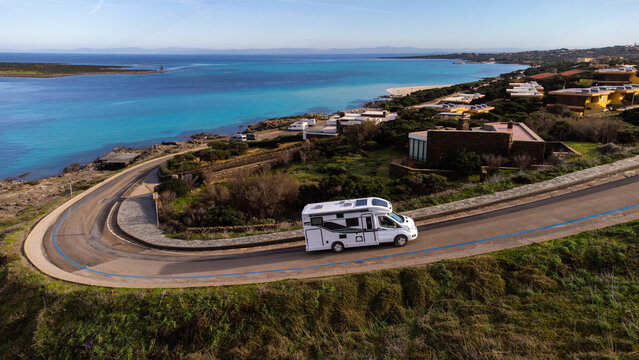 Aerial view of a white motorhome driving along a winding coastal road near the turquoise waters of La Pelosa beach in Stintino, Sardinia, Italy.