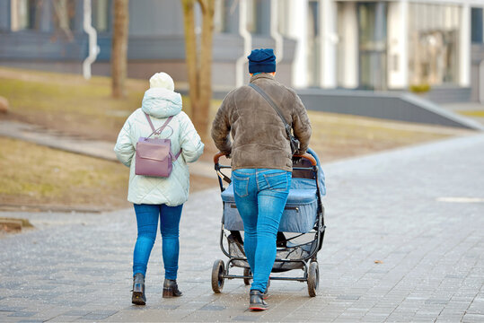 Parents in warm winter jackets and beanies pushing baby double stroller for twins together along paved city sidewalk covered with rock salt crystals. Young family walking together through winter city