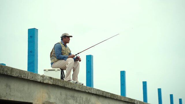 Happy senior man sitting by the sea and fishing with a rod after retirement. Active aging, healthy longevity, outdoor leisure, fulfilling retirement. Life insurance and financial security concept.
