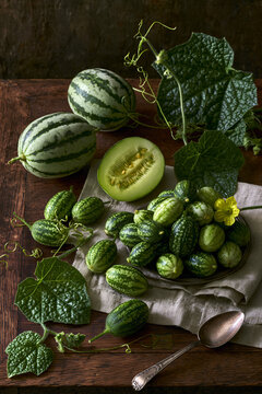 An elevated deconstructed botanical still-life featuring a harvest of small striped cucamelons, cut open melon fruits, leaves, and a vintage spoon.