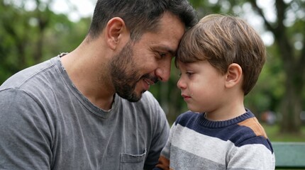 Fototapeta premium Father touches foreheads with his crying son in a park. Scene conveys empathy, emotional support, and fatherly love. Ideal for mental health and parenting articles.
