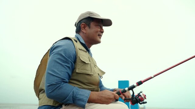 Happy senior man sitting by the sea and fishing with a rod after retirement. Active aging, healthy longevity, outdoor leisure, fulfilling retirement. Life insurance and financial security concept.
