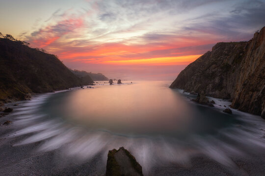 View of Silence Beach at sunset with long exposure waves washing over the pebble shore between rocky cliffs in Asturias, Spain.
