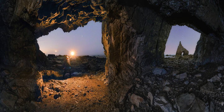 View of a person holding a flashlight inside a sea cave at Playa de Campiechos, looking out towards a rocky sea stack on the coast of Asturias, Spain.