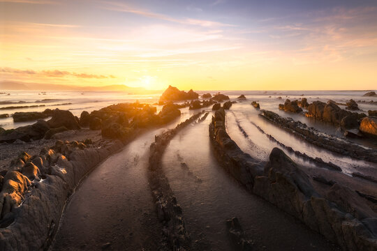 View of the unique flysch rock formations at Playa de Barrika during sunset with calm water reflecting the golden light in Elexalde, Basque Country, Spain.