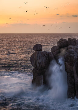 View of the Cantis de Papel rock formations with crashing waves and seagulls flying against a golden sunset sky in Moras, Lugo, Spain.