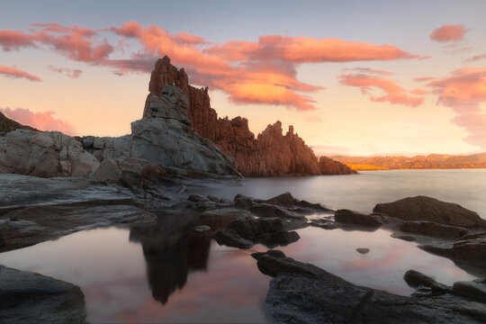 View of the iconic red granite Rocce Rosse rock formations reflecting in calm coastal tide pools at sunset in Arbatax, Sardinia, Italy.