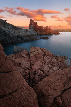 View of the iconic red granite rock formations of Rocce Rosse at sunset with calm sea and orange clouds in the sky in Arbatax, Sardinia, Italy.