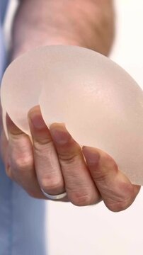 A plastic surgeon holding a silicone breast implant in hands, demonstrating flexibility and texture during a medical consultation in a clinic, healthcare and aesthetics industry, medical education.