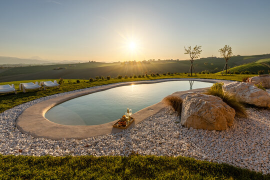 View of a natural-style swimming pool with a tray of wine and snacks, surrounded by white gravel, lush green grass, and rolling hills at sunset in Cupramontana, Marche, Italy.
