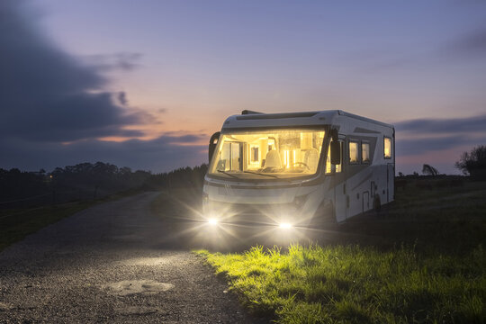 View of a modern camper van parked on a rural road at dusk with its headlights and interior lights illuminated in Portezuelo, Extremadura, Spain.