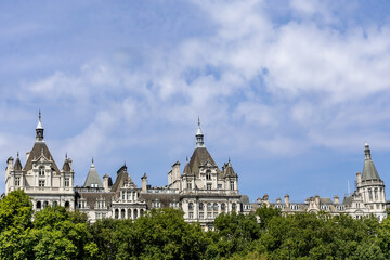 Obraz premium Historic Whitehall Court buildings rise above leafy trees along the River Thames in Westminster, London, England, featuring ornate Victorian architecture beneath a blue and cloudy summer sky