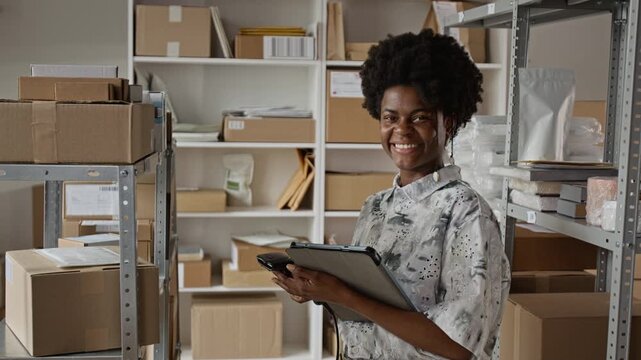 Portrait of young adult African American woman holding tablet and barcode scanner in warehouse, turning to smile at camera. Perfect for e-commerce logistics and small business shipping promos
