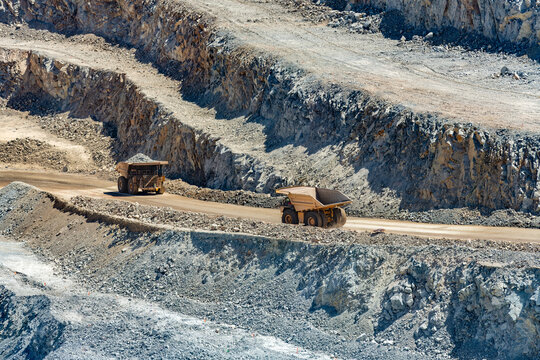 Large industrial mining trucks transport heavy copper ore along a dirt haul road cut into the rocky terraced walls of a massive open pit mine in Chile.