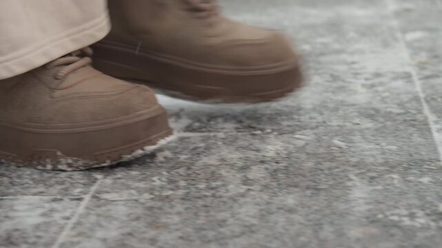 Low-angle close-up of two muddy brown boots walking on wet dark gray tile or stone, captured midstep with heel-to-toe roll, mud clinging to soles, and small water splashes along grout lines; diffuse