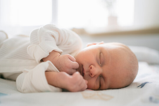 Newborn baby sleeping on a white blanket with closed eyes, hands resting near the face, feeling comfortable and safe in a warm environment, representing early childhood and tender moments