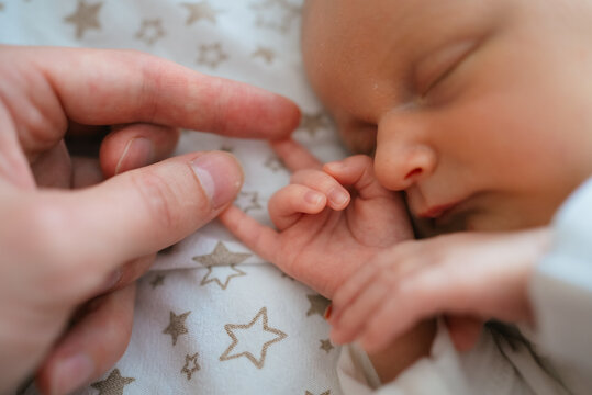 Newborn baby closes eyes while gently gripping an adult's finger, showcasing a tender moment of connection, love, and protection within the family unit