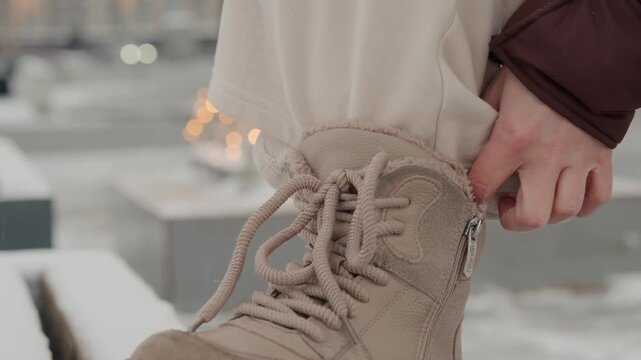Close low angle view of person securing beige winter boot on snowy ledge while hand pulls laces, smooths padded collar and settles side closure during cold weather dressing, with boot, lace, hand