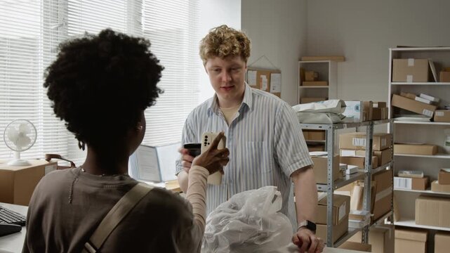Young adult Caucasian man working at e-commerce pickup point inspecting apparel returned by Black woman customer. Clip represents retail returns, order fulfillment, and customer service