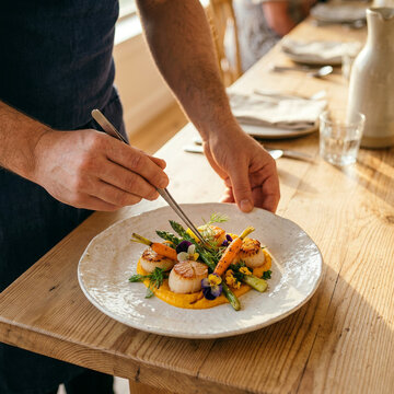 Chef carefully plating a vegetable and flower accented dish on a white ceramic plate in warm dining