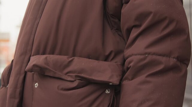 Torso and arm in dark brown padded coat with hand sliding into pocket, wet droplets and damp patches on sleeve and pocket, cool urban outdoor light, winter commute mood, with coat, pocket, hand