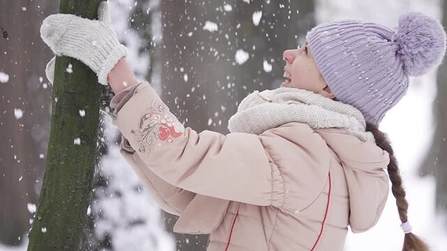Slow motion. Teen girl shakes a young tree from snow in a winter park.