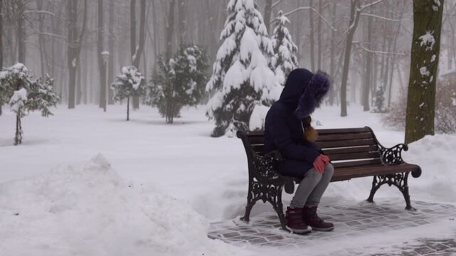 Beautiful teen girl with long hair in a dark jacket with a hood sitting on a bench in the winter snow-covered park.
