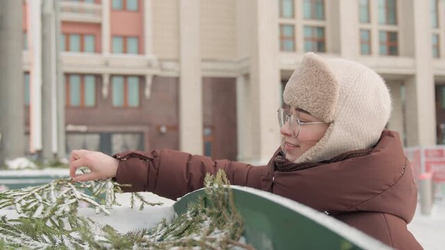 Elderly woman leans over low metal railing, carefully straightening evergreen wreath with gloved hands in snowy courtyard, creating quiet seasonal decoration beside residential building