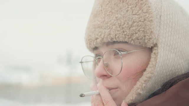Close head and shoulder view of young woman wearing fur lined earflap hat and round glasses outdoors in snowy overcast weather, visible breath and calm thoughtful expression, with woman, female