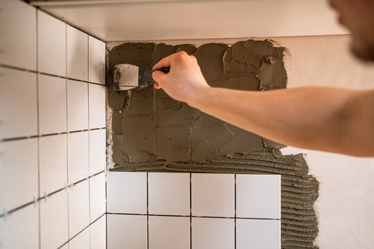 Worker spreading cement adhesive with a trowel while installing modern ceramic backsplash tiles in a kitchen during a home renovation, close-up on hands and wall progress