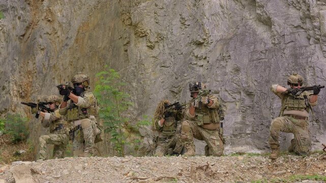 A special forces team conducts a military operation, with soldiers aiming rifles from defensive positions against a rugged rock face background.