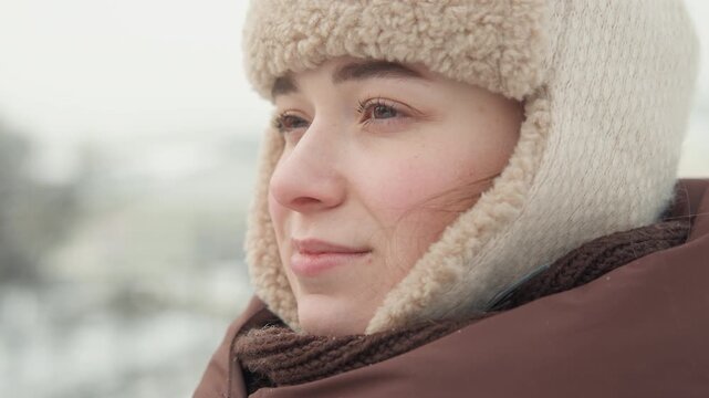 Close-up motion portrait of young person in shearling hat with earflaps and padded jacket, using both hands to adjust metal-rimmed glasses before turning toward snowy distance in soft daylight