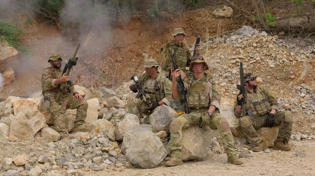 An elite team of special forces soldiers in full tactical gear pose together with their rifles amidst smoke and rocks during a military operation in a rugged combat zone.