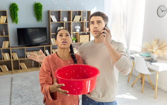 Couple in apartment catch ceiling leak with bucket. Water drips as the man calls a plumber on phone, while homeowners use a bucket to limit damage inside. Clear home repair emergency concept.