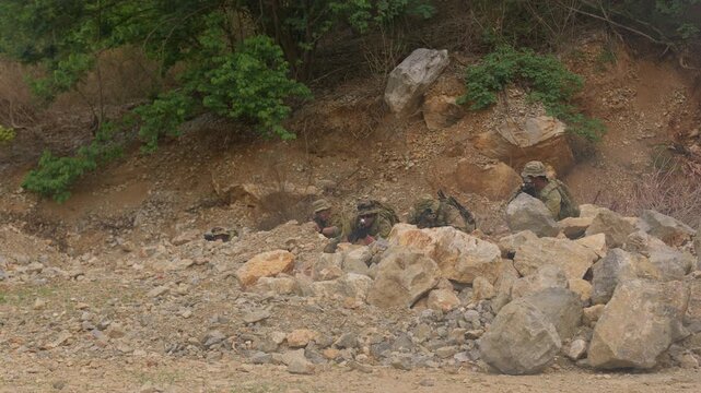 A military squad in camouflage takes a defensive position, aiming rifles from behind rocky cover during a reconnaissance mission or tactical training exercise.