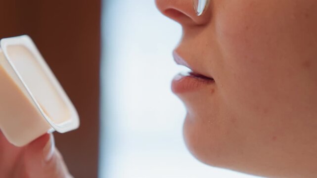 Cropped side-profile view shows adult with glasses holding small plastic container of sauce near mouth, rim tilted toward pursed lips in calm indoor tasting moment with soft focus and intimate detail