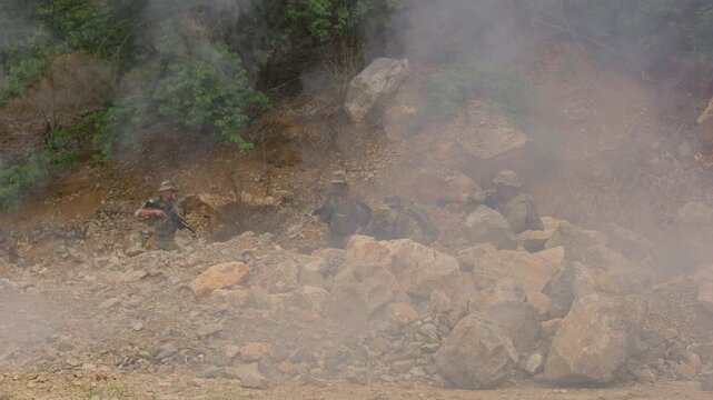 A military squad in camouflage takes a defensive position, aiming rifles from behind rocky cover during a reconnaissance mission or tactical training exercise.