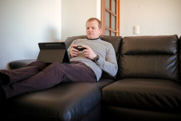 Fototapeta premium Man relaxing on couch playing video game with tablet and controller