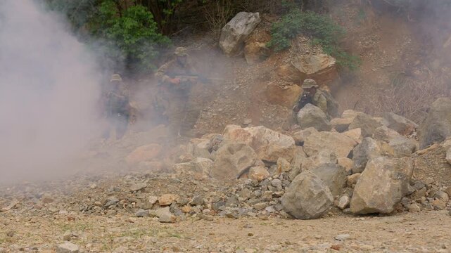 A military squad conducts a tactical operation, advancing through heavy smoke while using the rocky, hostile terrain for cover during a simulated combat mission.