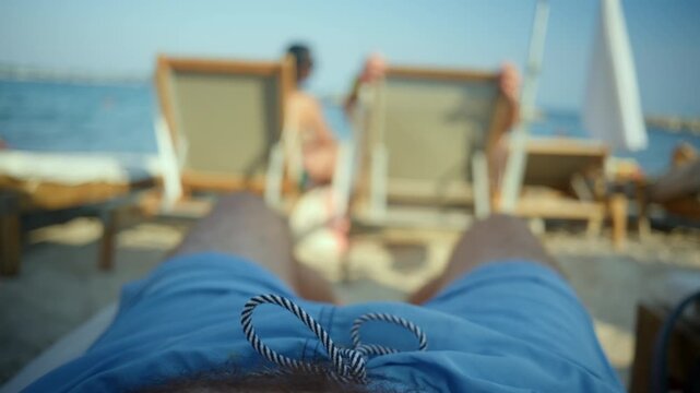Person's POV lying on a beach chair, wearing blue shorts, enjoying the sun and blurred people in the background