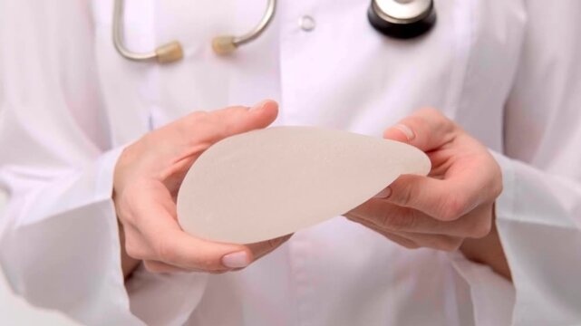 Doctor in a medical coat holding a round breast implant during a consultation, healthcare advertising, plastic surgery, aesthetic medicine.