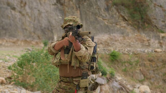 A special forces soldier in full tactical gear aims a sniper rifle with a suppressor, conducting a combat mission in a rugged mountain environment.