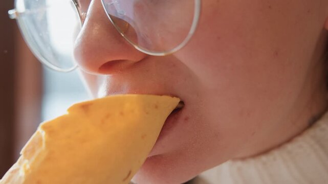 Close-up side profile of young person wearing round glasses biting wrapped sandwich or tortilla indoors, with visible lip contact, small crumb on chin, soft natural light, casual eating scene